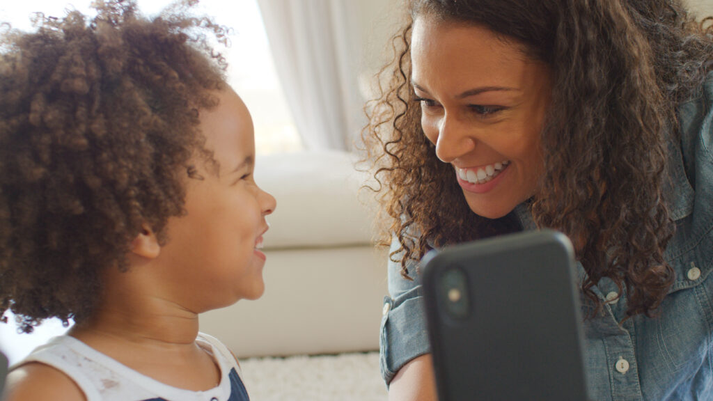 Woman holding a phone up while she smiles at a child who is smiling at her.