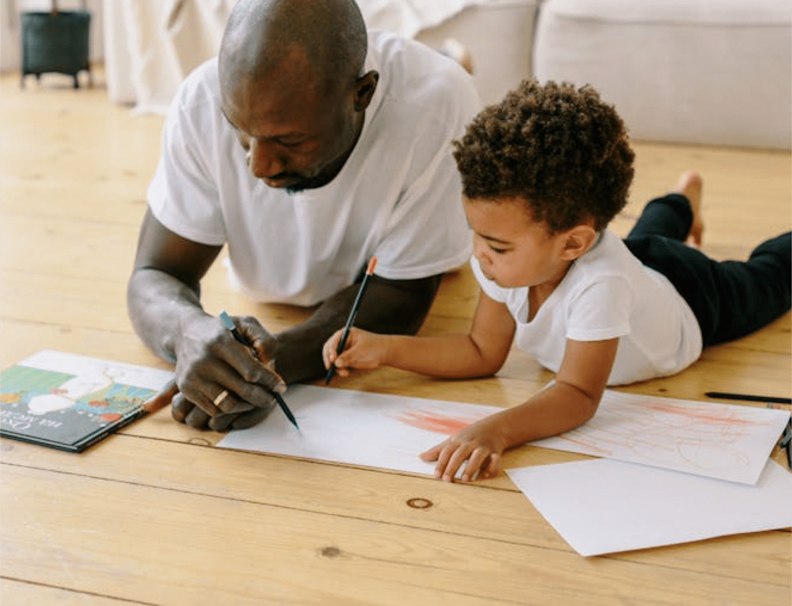 Child and father laying on floor coloring while looking at a book.