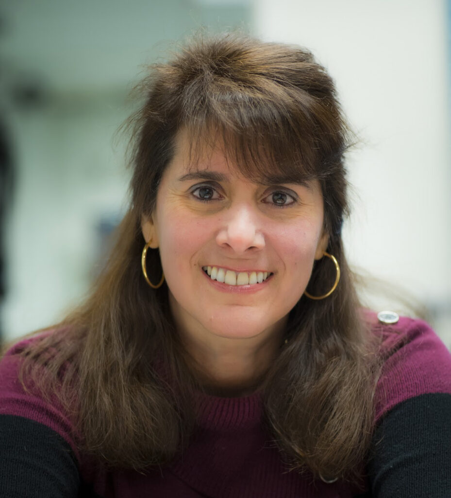 Liz has long brown hair, bangs, and is smiling at the camera.